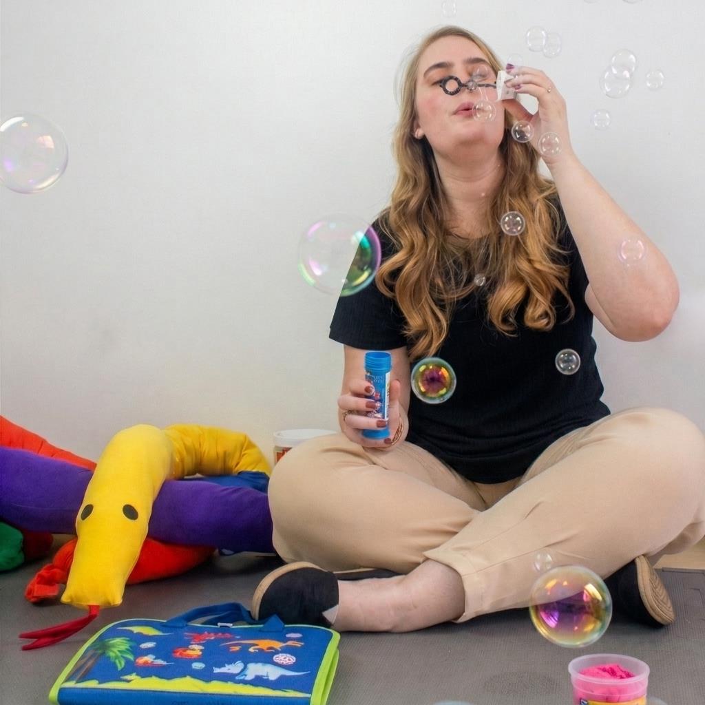 Letícia Neufeld soprando bolhas de sabão cercada por brinquedos sensoriais coloridos no chão