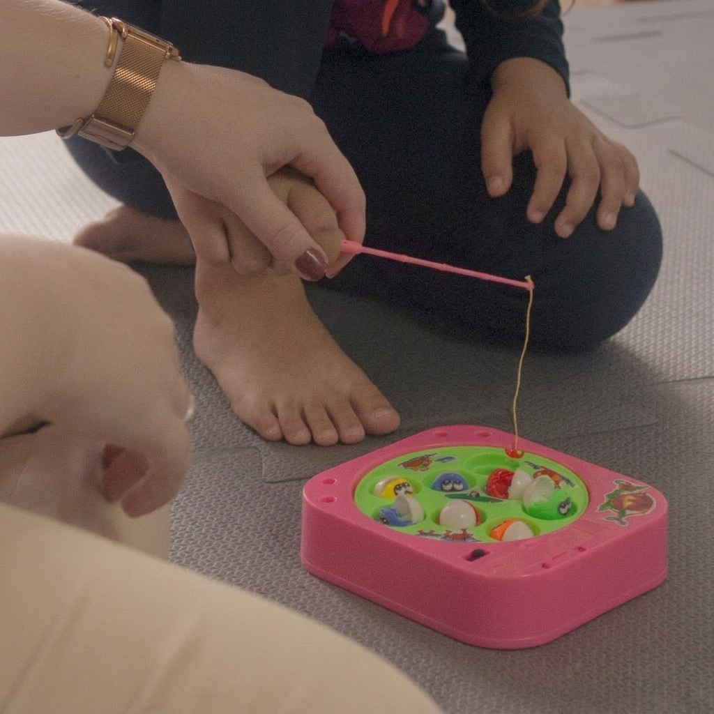 Mão de terapeuta com relógio dourado auxiliando criança a segurar varinha de pesca rosa em jogo de pescaria infantil no tatame